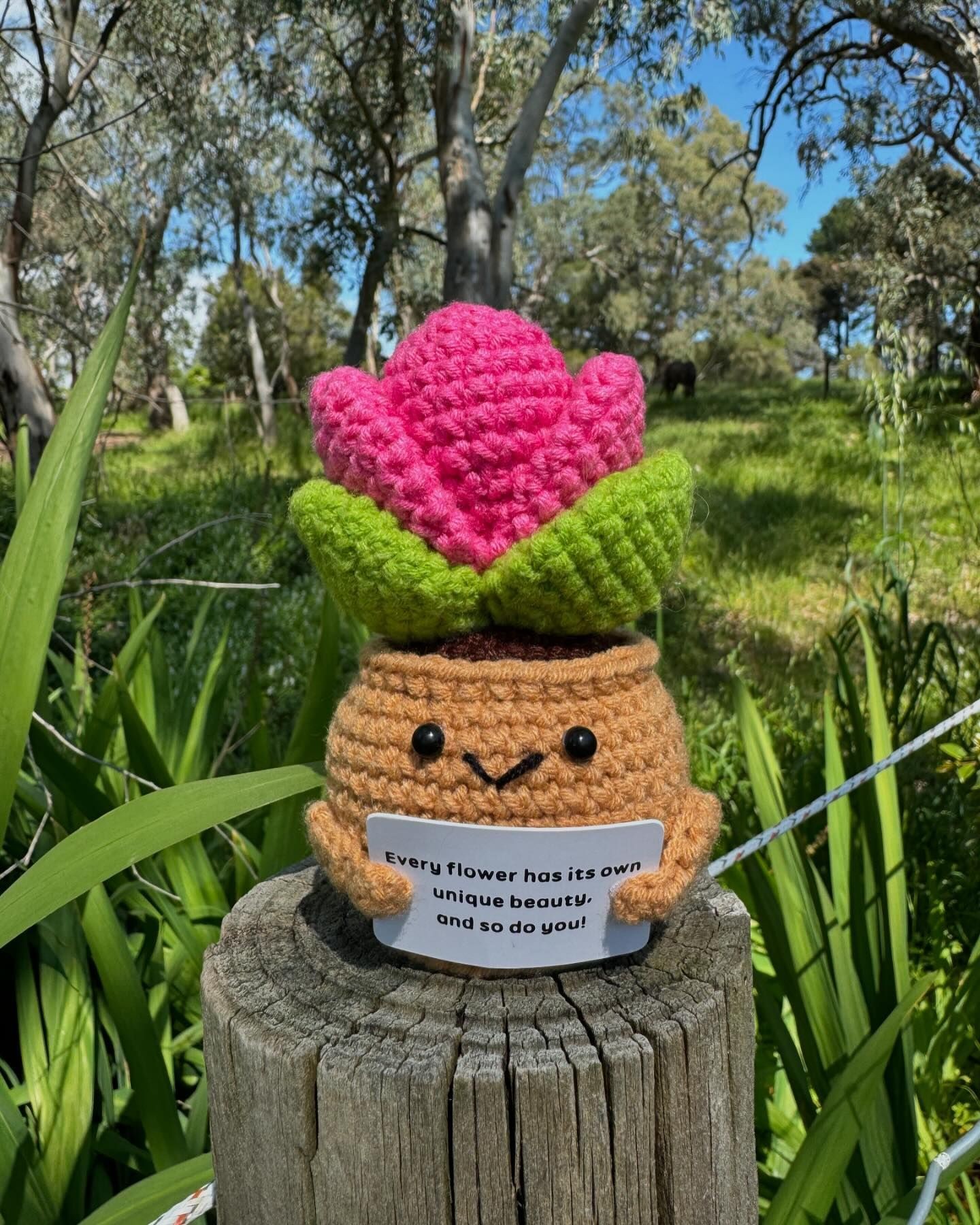 Crocheted flower plush toy holding sign, outdoors with greenery background.